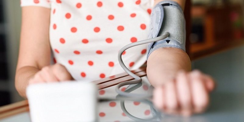 Bluthochdruck_web_800.jpg Eine Person mit einem Blutdruckmessgerät am Arm, das auf einem Tisch liegt. Im Hintergrund ein unscharfer Raum.