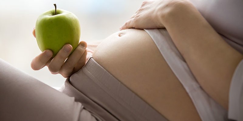 Ernaehrung-Schwangerschaft_Web800-800×400.jpg Eine schwangere Person hält einen grünen Apfel und berührt sanft ihren Bauch.