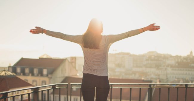 Gesundes_Immunsystem.jpg Eine Person mit ausgestreckten Armen steht auf einem Balkon und blickt in die Sonne über einer Stadt.