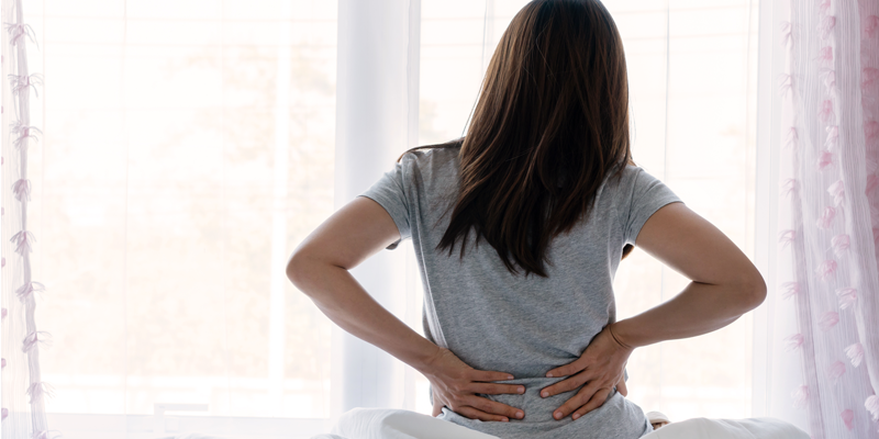 Ruecken_web.png Eine Person mit langen Haaren sitzt auf einem Bett und hält sich den Rücken vor einem Fenster.