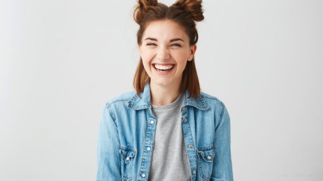Lachende junge Frau mit Duttfrisur, trägt eine Jeansjacke über einem grauen T-Shirt, vor einem hellen Hintergrund.