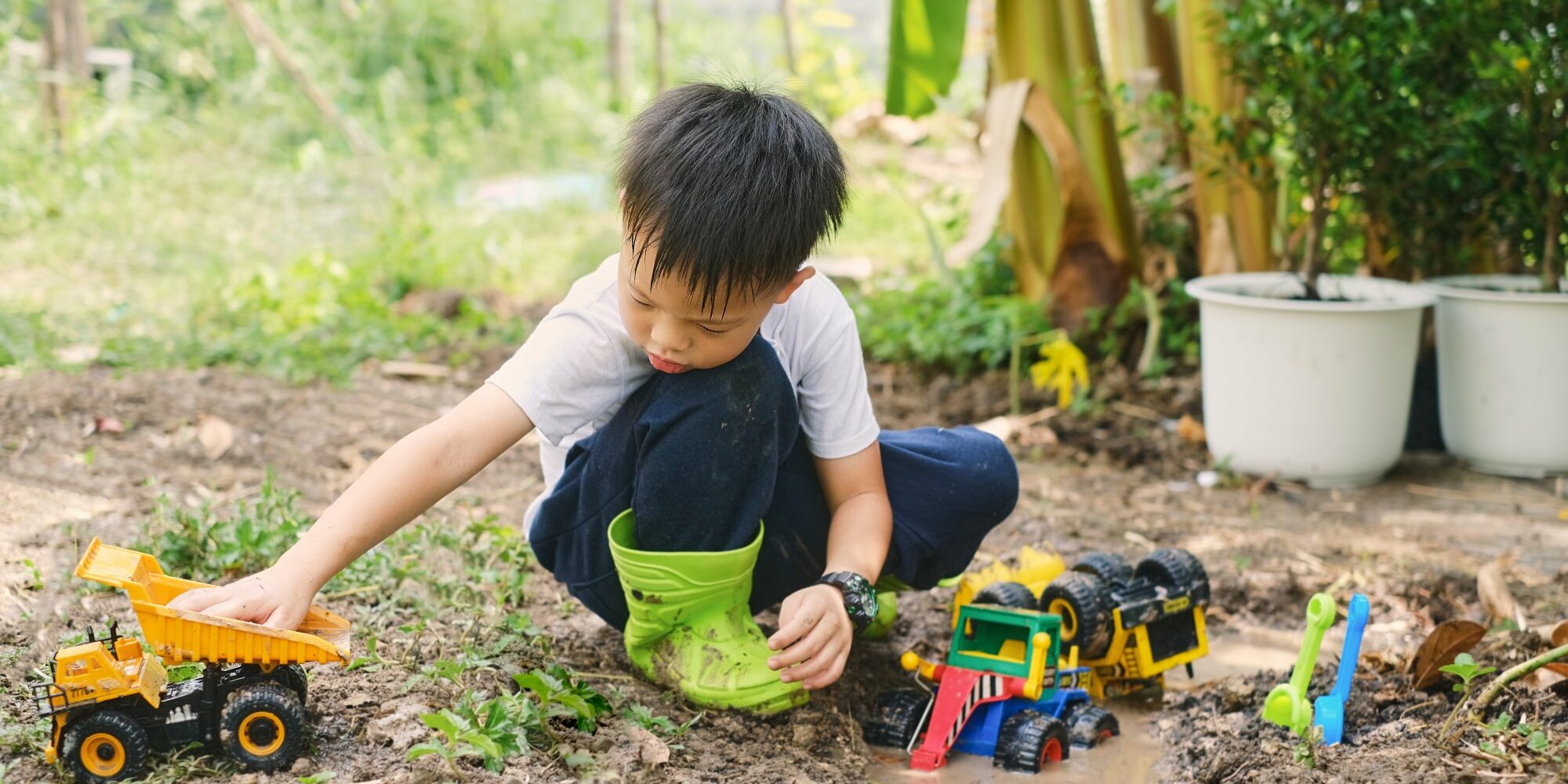 Ein Junge im Garten spielt mit verschiedenen Spielzeugfahrzeugen, darunter ein Lkw und ein Bagger, während er in matschigem Boden sitzt.