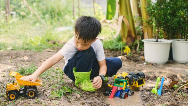 Ein Junge im Garten spielt mit verschiedenen Spielzeugfahrzeugen, darunter ein Lkw und ein Bagger, während er in matschigem Boden sitzt.