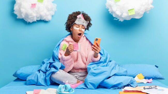 Eine Person in einem blauen Bett, umhüllt von einer Decke, schaut müde auf ihr Handy und hält eine Tasse in der Hand. Wolken mit Haftnotizen im Hintergrund.