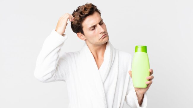 Ein Mann in einem weißen Bademantel hält eine grüne Shampoo-Flasche und sieht nachdenklich aus.