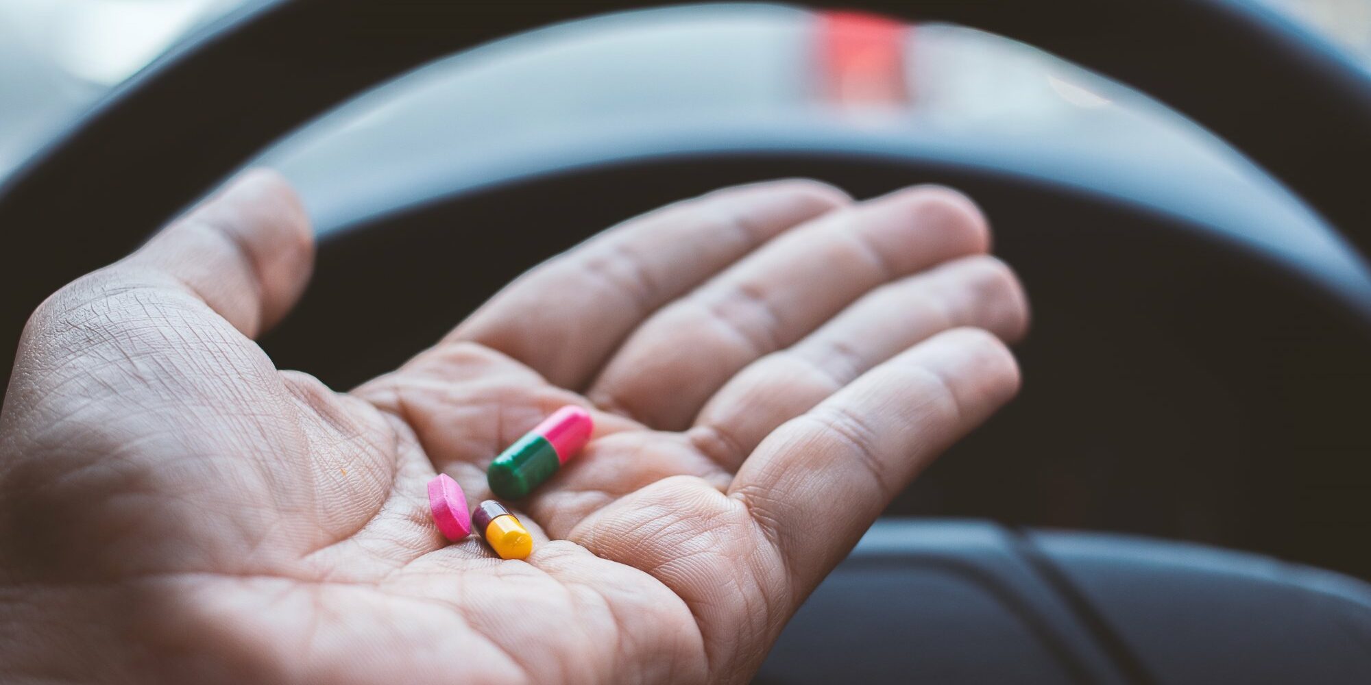 Eine Hand hält bunte Kapseln über dem Lenkrad eines Fahrzeugs.