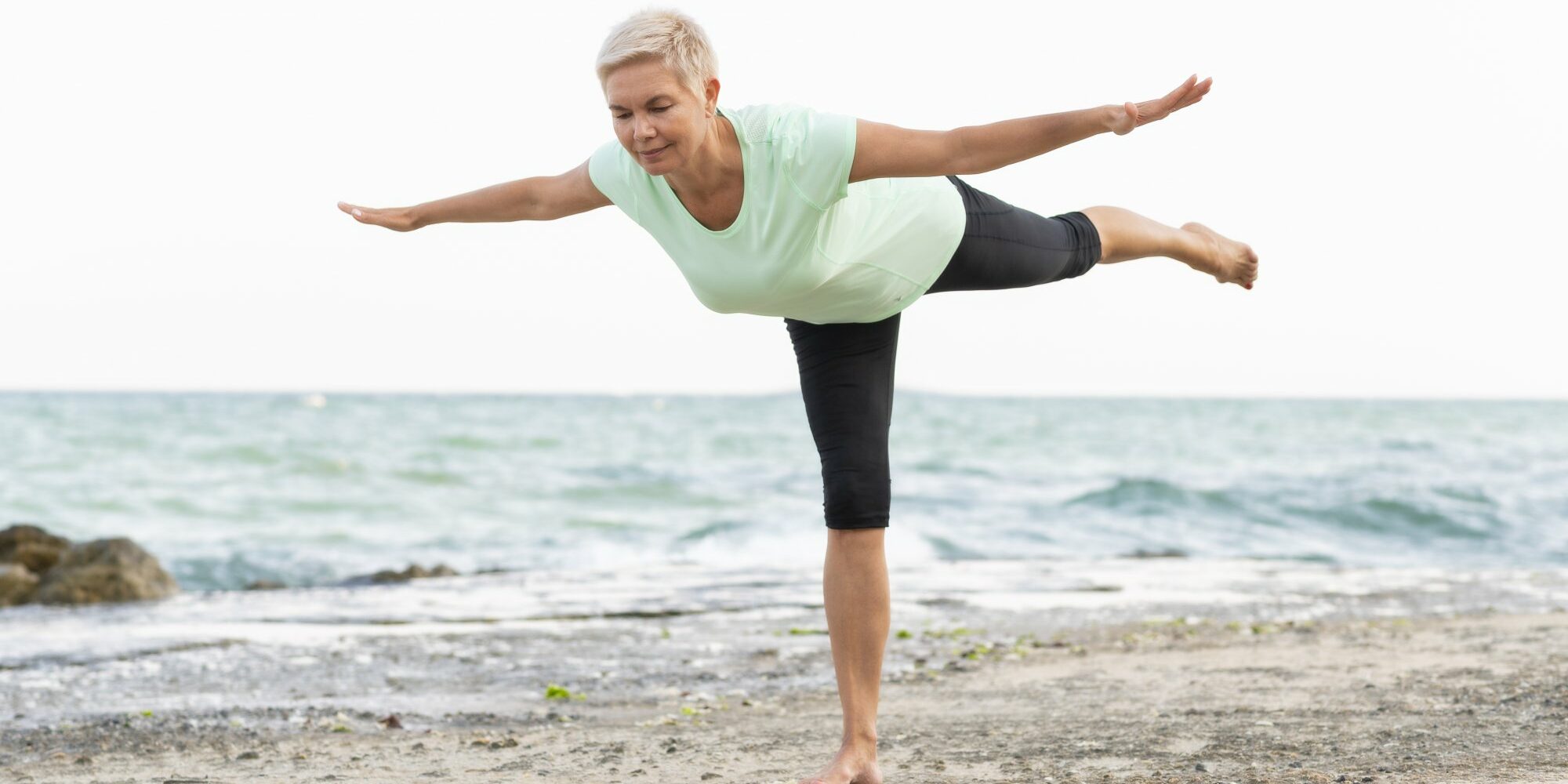 Eine Frau in einem hellgrünen T-Shirt und schwarzen Leggings balanciert auf einem Bein am Strand.