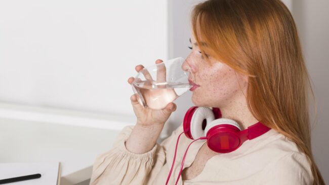 fluessigkeitsmangel.jpg Eine Person mit roten Kopfhörern trinkt aus einem Glas Wasser und sitzt an einem Schreibtisch.