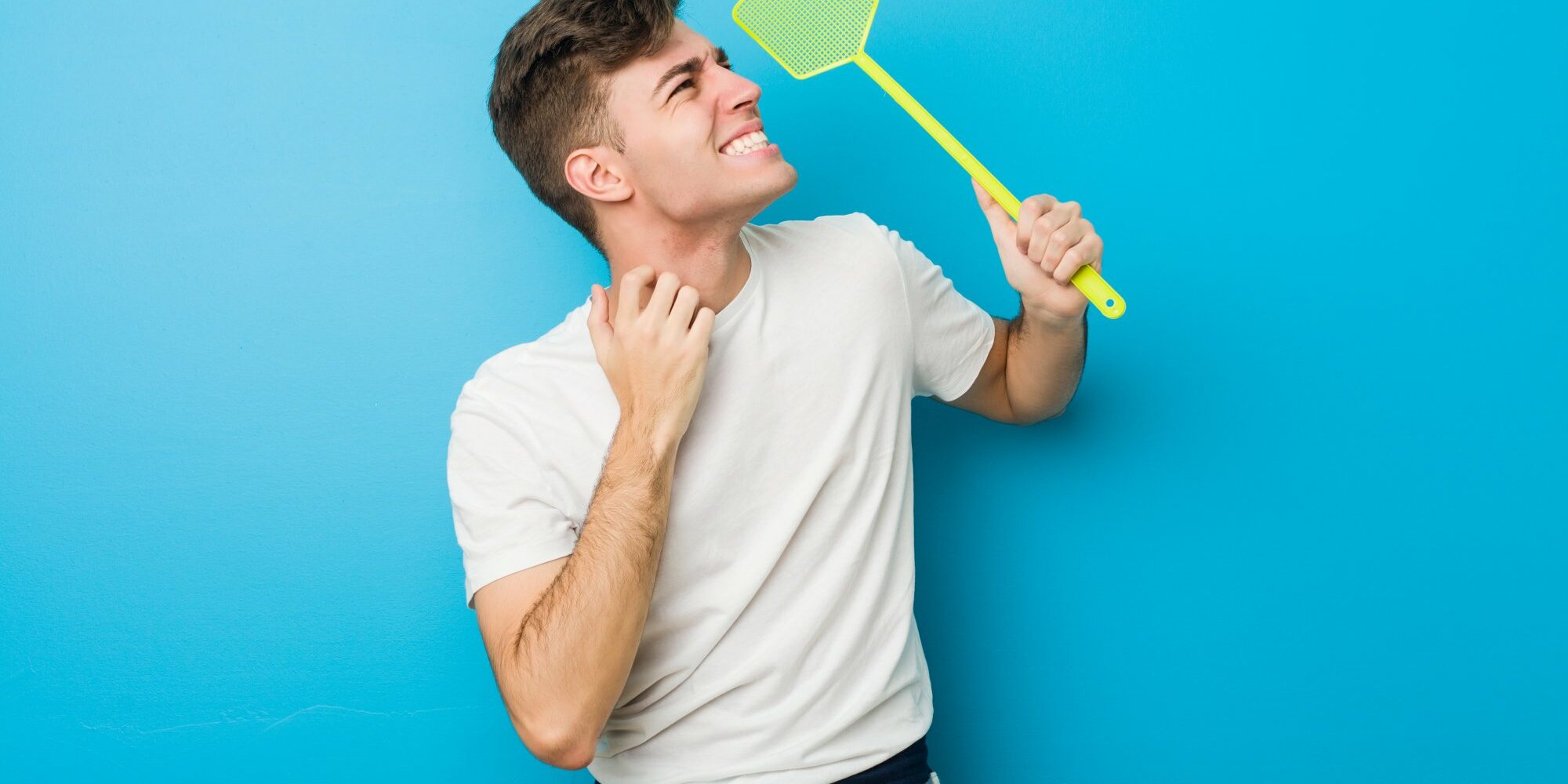 plagegeister.jpg Junger Mann in einem weißen T-Shirt hält ein grünes Insektenspray mit einem lächelnden Gesicht vor einer blauen Wand.
