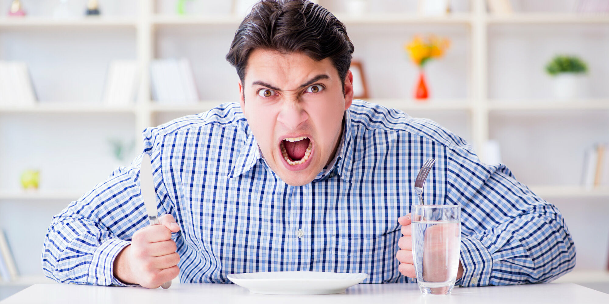 Hangry.jpg Ein wütender Mann hält ein Messer und eine Gabel über einem leeren Teller, während er an einem Tisch sitzt.
