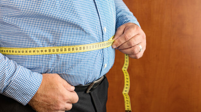 Mann misst seinen Bauchumfang mit einem gelben Maßband, trägt ein kariertes Hemd und dunkle Hose.