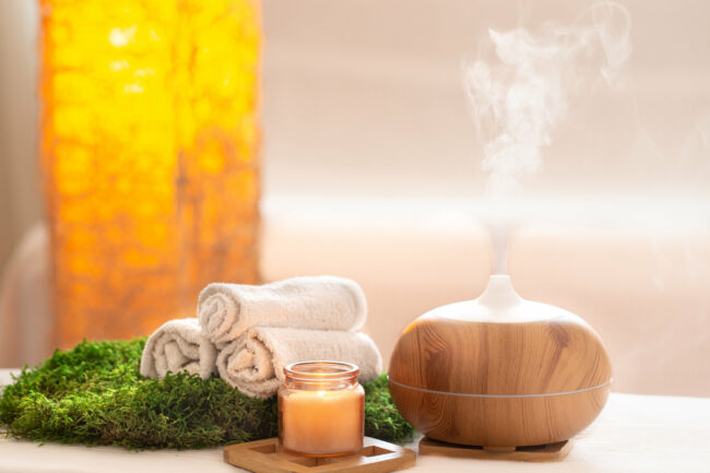 aromatherapie.jpg Ein Aromadiffusor aus Holz mit aufsteigendem Dampf, umgeben von Handtüchern, einer Kerze und grünem Moos.