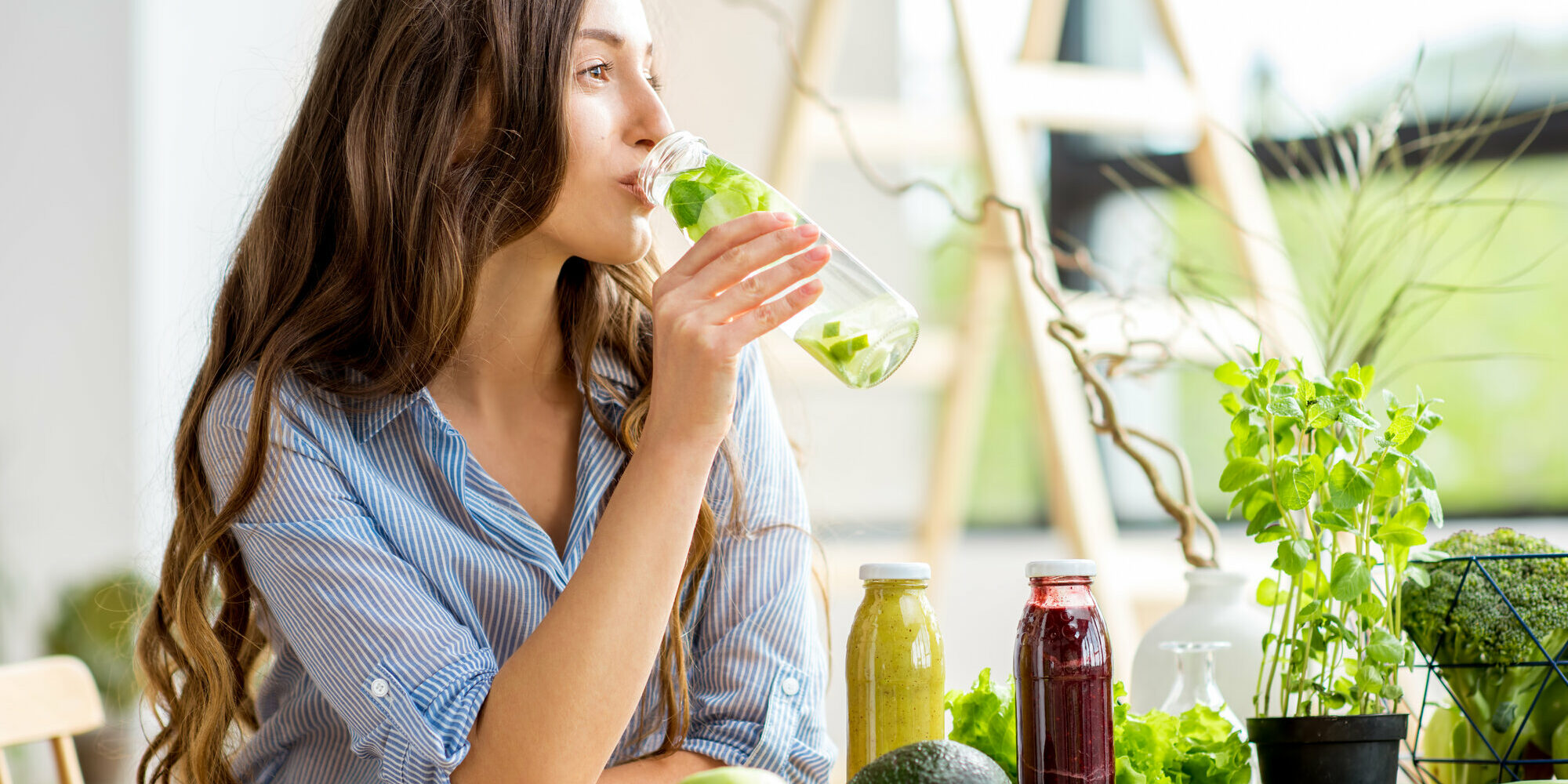 Eine Frau trinkt aus einer Glasflasche mit frischer Minze, umgeben von gesunden Lebensmitteln und Pflanzen.