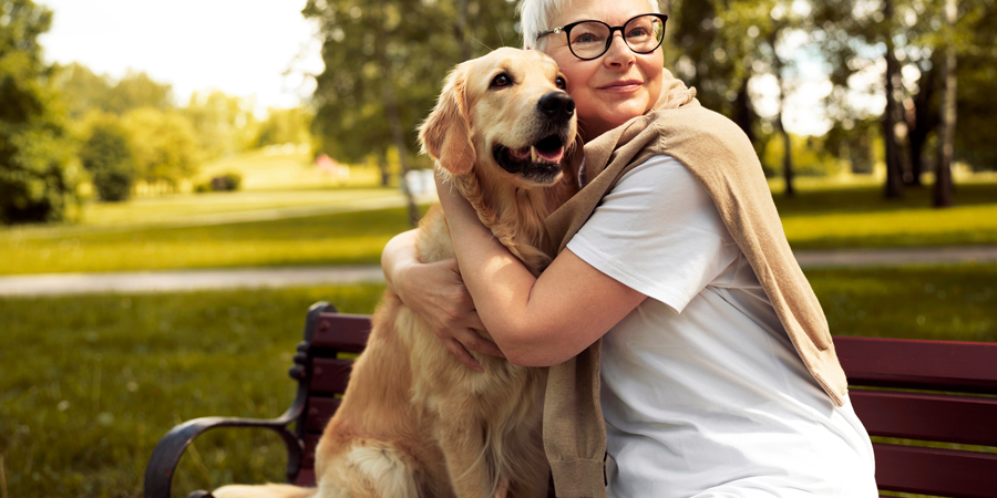 Eine Frau mit kurzen, blonden Haaren und Brille umarmt einen golden retriever auf einer Bank im Park.