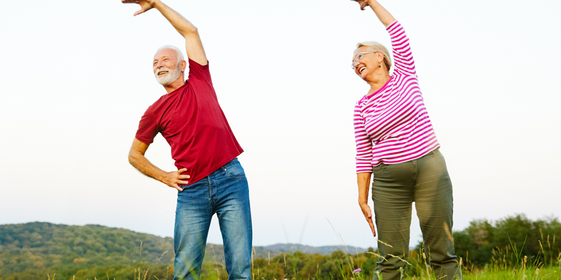Ein älteres Paar führt Dehnübungen im Freien auf einer Wiese durch, umgeben von Landschaft und Hügeln.