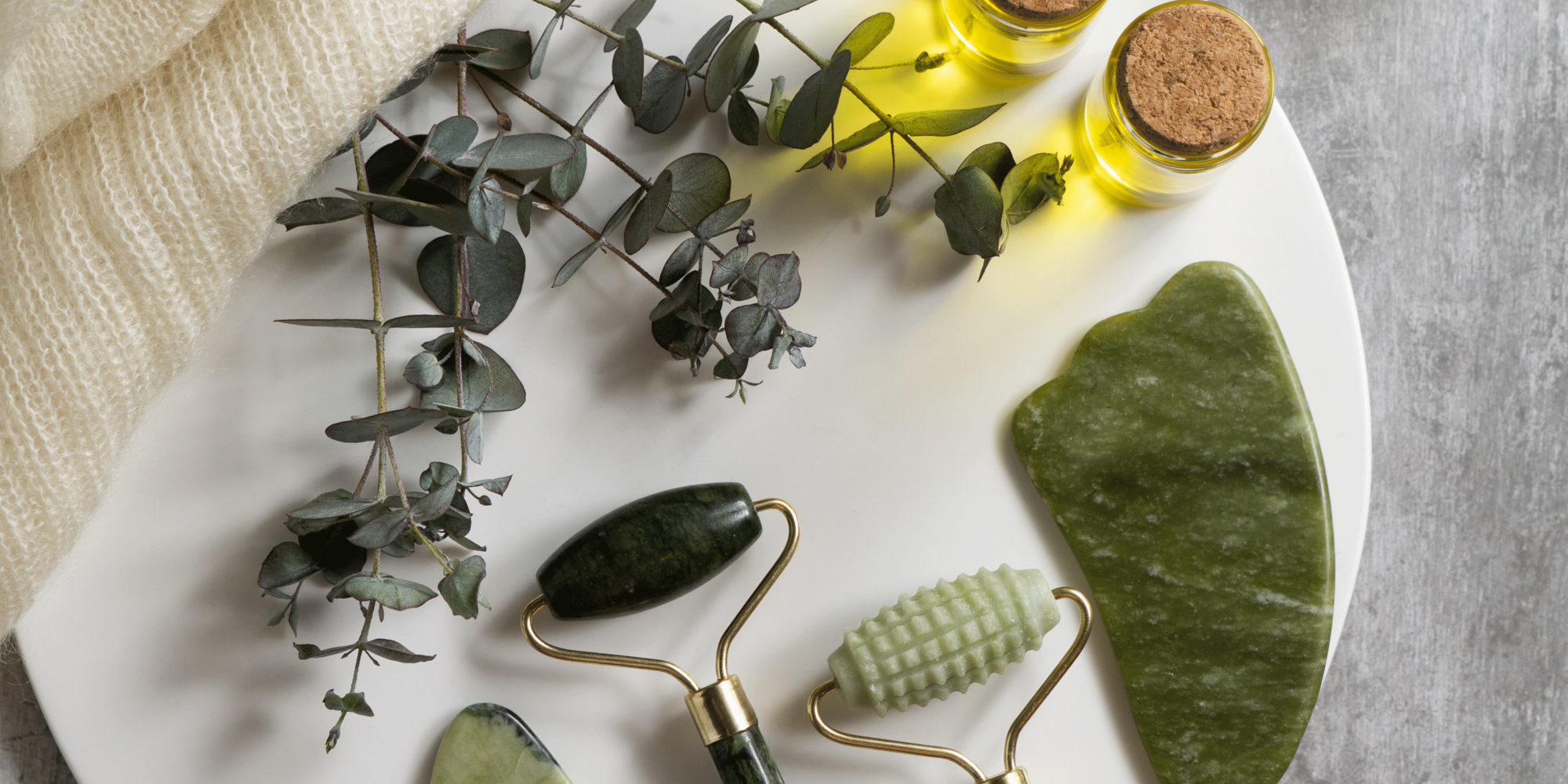 Ein weißer Teller mit drei Jade-Rollern, einem gua sha Stein, zwei Fläschchen ätherischem Öl und Eukalyptuszweigen.