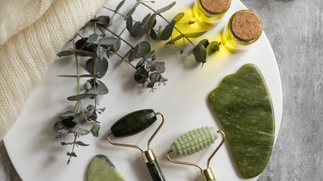 Ein weißer Teller mit drei Jade-Rollern, einem gua sha Stein, zwei Fläschchen ätherischem Öl und Eukalyptuszweigen.