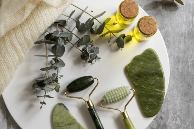 nachhaltige-kosmetik.png Ein weißer Teller mit drei Jade-Rollern, einem gua sha Stein, zwei Fläschchen ätherischem Öl und Eukalyptuszweigen.