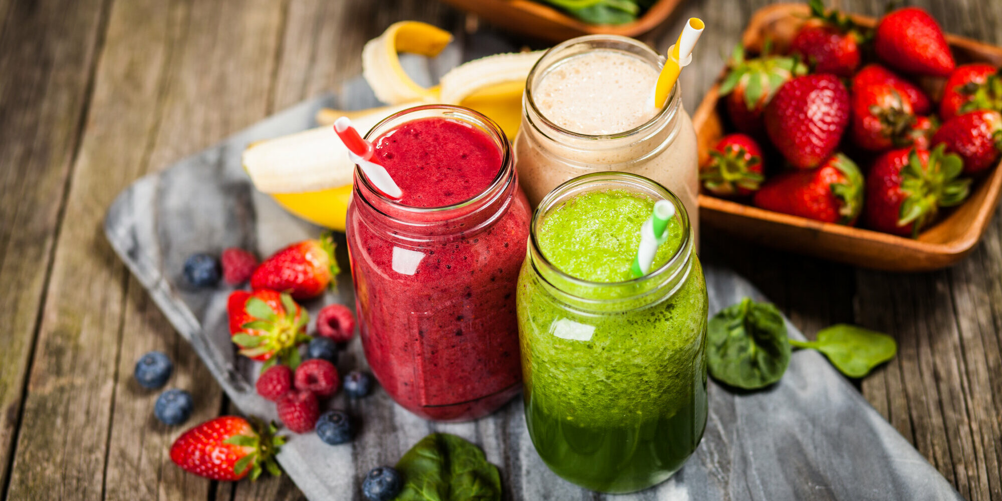 Drei bunte Smoothies in Gläsern mit Strohhalmen, umgeben von frischen Früchten und Spinat auf einer Holzunterlage.
