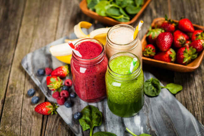 Smoothies__2_.jpeg Drei bunte Smoothies in Gläsern mit Strohhalmen, umgeben von frischen Früchten und Spinat auf einer Holzunterlage.