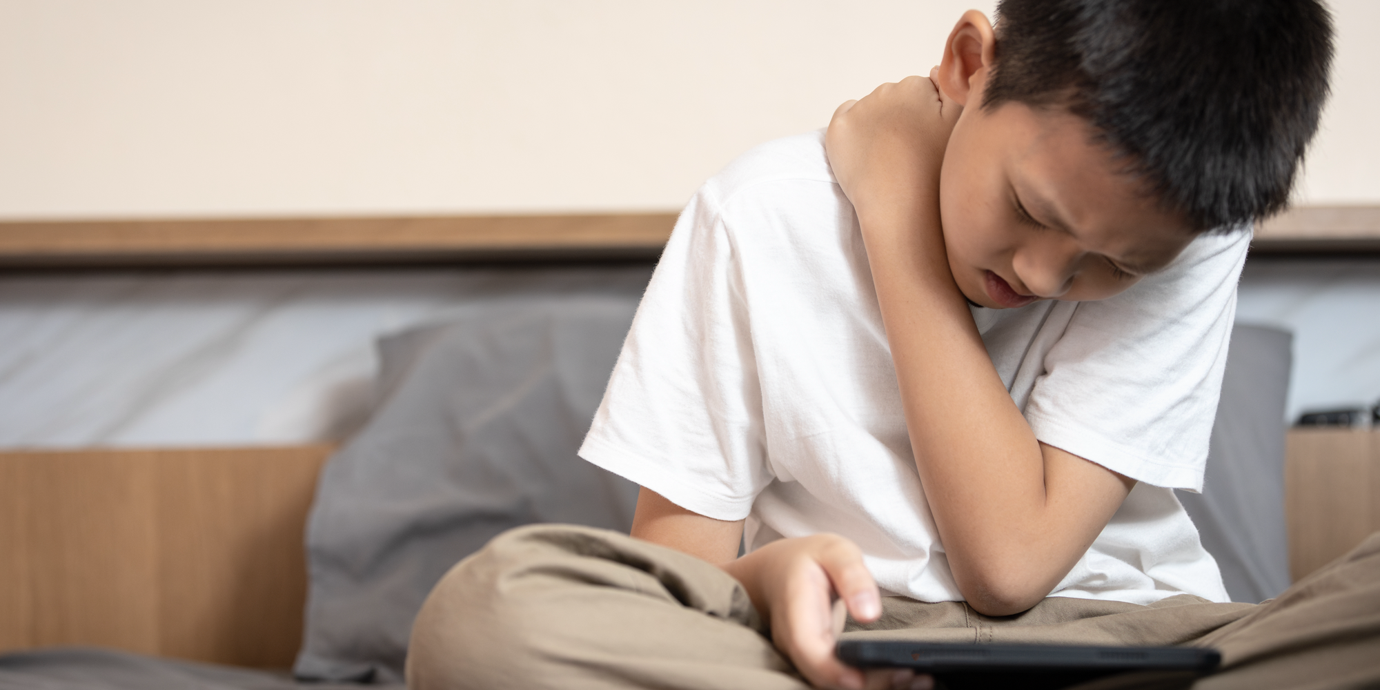 rueckenfit.png Ein Junge sitzt auf einem Bett, hält ein Tablet und fasst sich schmerzverzerrt an den Hals.