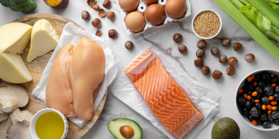 Proteinreiche_Ernaehrung.png Verschiedene Lebensmittel wie Hähnchenbrust, Lachs, Käse, Eier, Avocado und Gemüse auf einem Tisch.