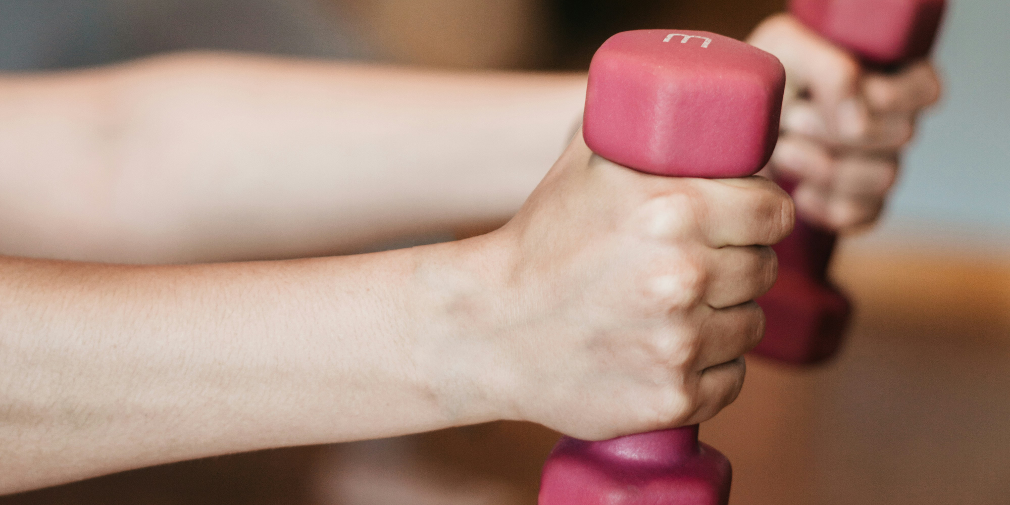 mobility-training.png Hände halten zwei pinkfarbene Kurzhanteln in einem Fitnessstudio.