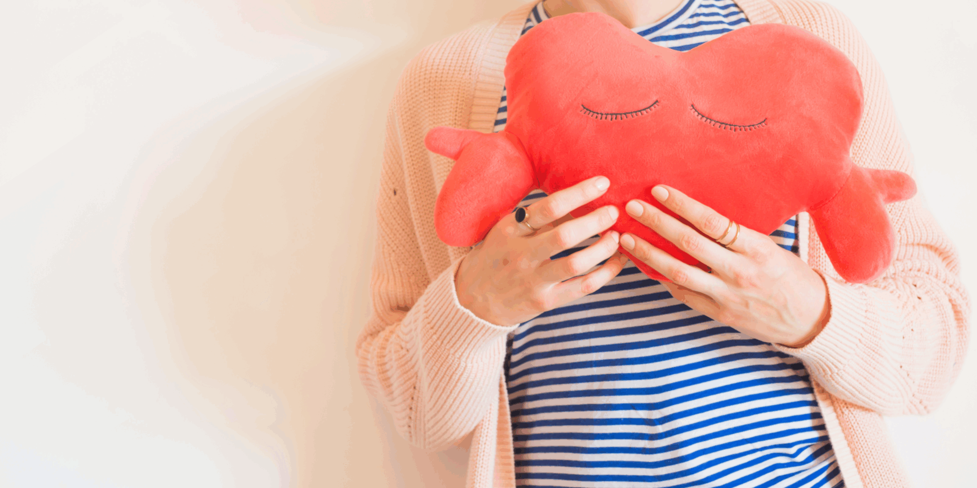 Eine Person hält ein rotes herzförmiges Kissen mit geschlossenen Augen, gekleidet in gestreiftes Oberteil und eine helle Strickjacke.