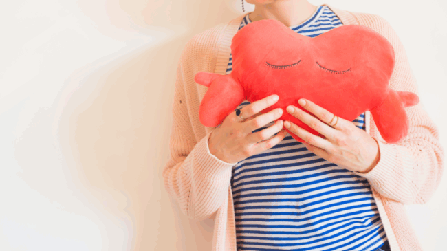 Eine Person hält ein rotes herzförmiges Kissen mit geschlossenen Augen, gekleidet in gestreiftes Oberteil und eine helle Strickjacke.
