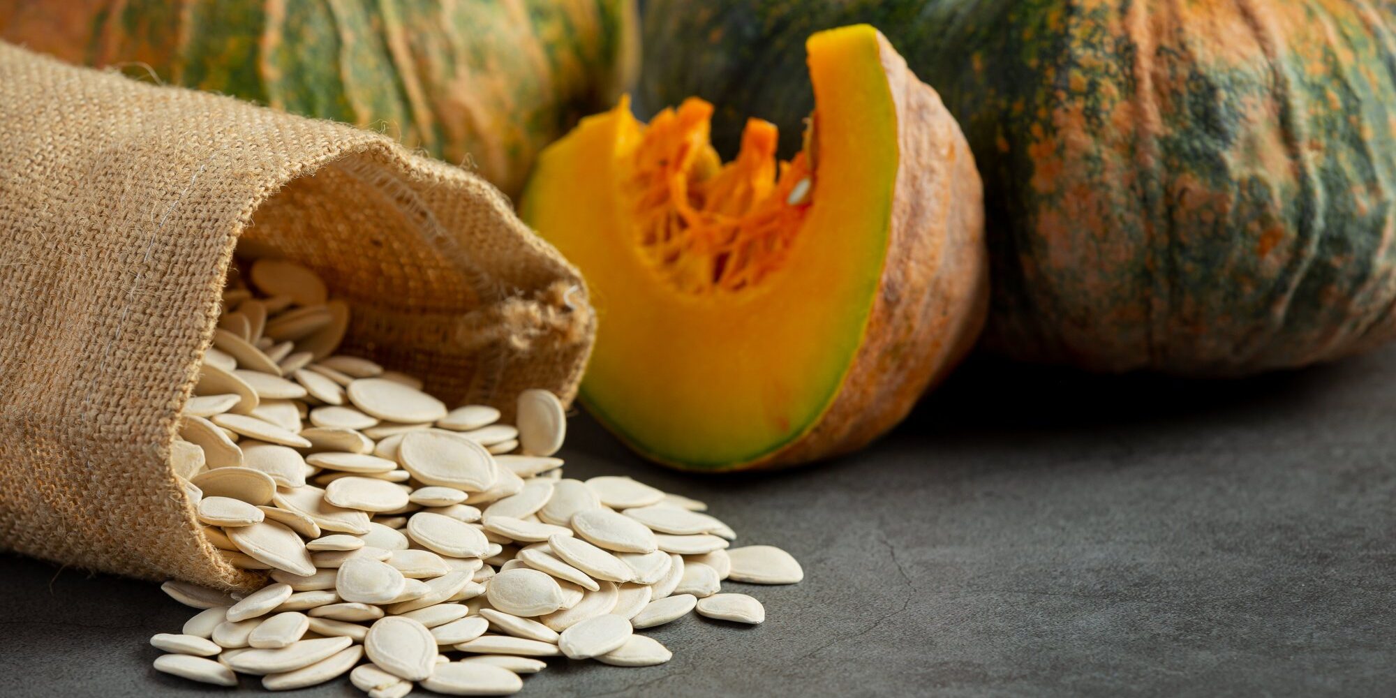 Kuerbis.jpg Eine geöffnete Jute-Tasche mit Kürbissamen und zwei Kürbissen, einer davon halbiert, auf einem dunklen Hintergrund.