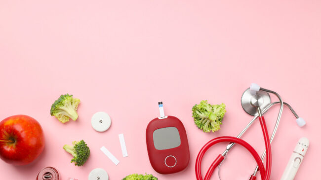 Gesunde Lebensmittel und medizinische Geräte auf rosa Hintergrund, darunter Apfel, Brokkoli, Blutzuckermessgerät und Stethoskop.