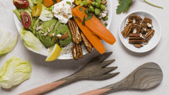 Bunte Salatschüssel mit Avocado, Karotten, Erbsen, Mais, Tomaten und Nüssen, begleitet von Holzbesteck und einer Schale Nüssen.