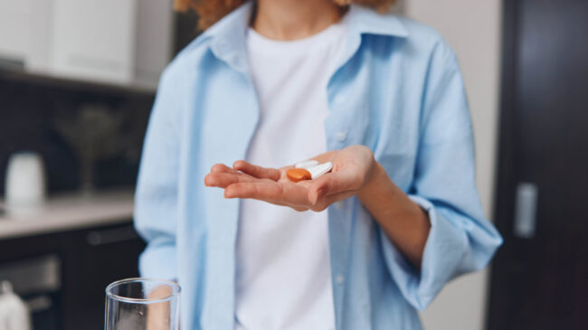 medikamenteneinnahme.jpeg Eine Person hält zwei Kapseln in einer Hand, während in der anderen ein Glas Wasser steht, in einer modernen Küche.