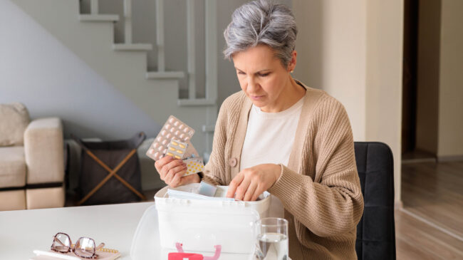 Eine Frau in einem hellen Raum untersucht einen durchsichtigen Medikamentenbehälter und hält eine Packung Tabletten.